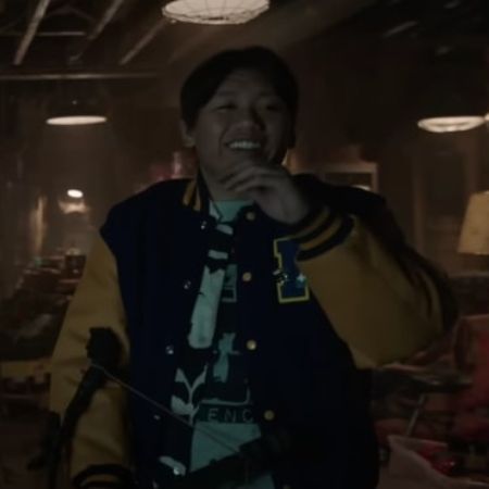Jacob Batalon is laughing, wearing a yellow and blue University jacket.